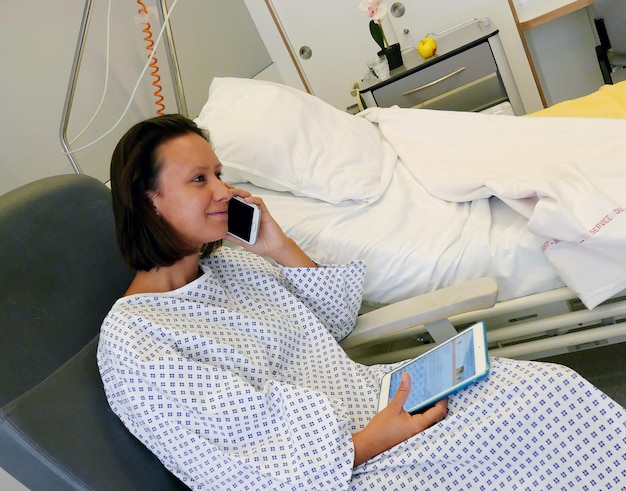 Photo young woman wearing hospital gown using mobile phone and digital tablet