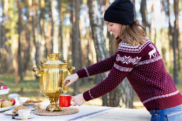 Young woman wearing jersey with hat pouring some tea using vintage russian teapot samovar