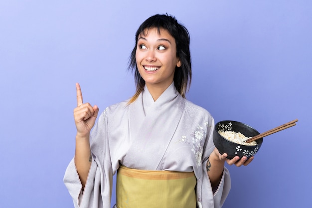 Giovane donna che indossa un kimono sul muro blu con l'intenzione di realizzare la soluzione sollevando un dito mentre si tiene una scodella di noodles con le bacchette