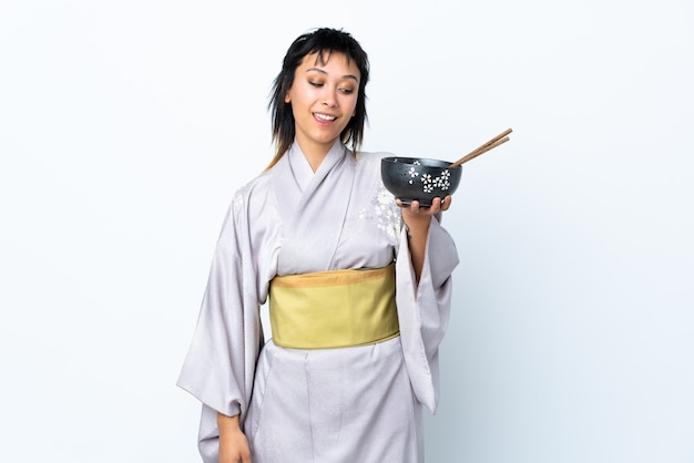 Young woman wearing kimono holding a bowl of noodles over isolated white background with happy expression