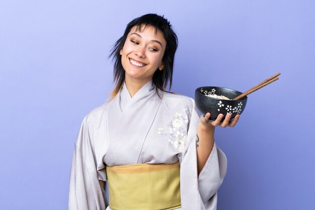 Young woman wearing kimono over isolated blue wall with happy expression while holding a bowl of noodles with chopsticks