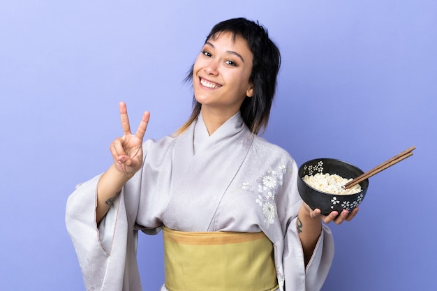写真 孤立した青い笑顔で着物を着た若い女性笑顔と箸で麺のボウルを押しながら勝利のサインを示す