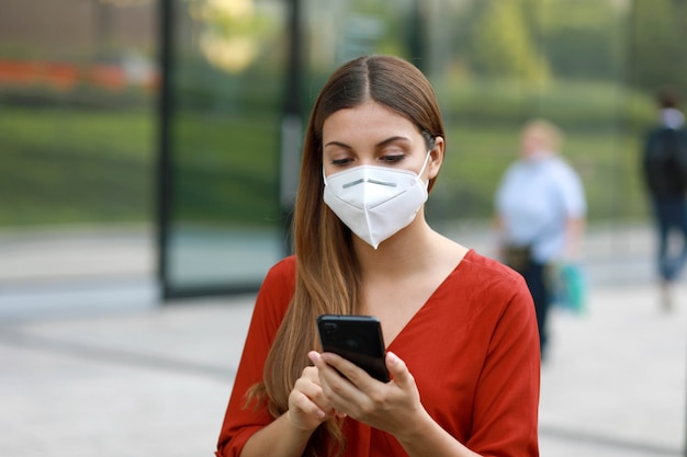 Giovane donna che indossa la maschera ffp2 kn95 utilizzando l'app del telefono cellulare in una strada cittadina per aiutare la ricerca dei contatti e l'autodiagnostica in risposta al coronavirus