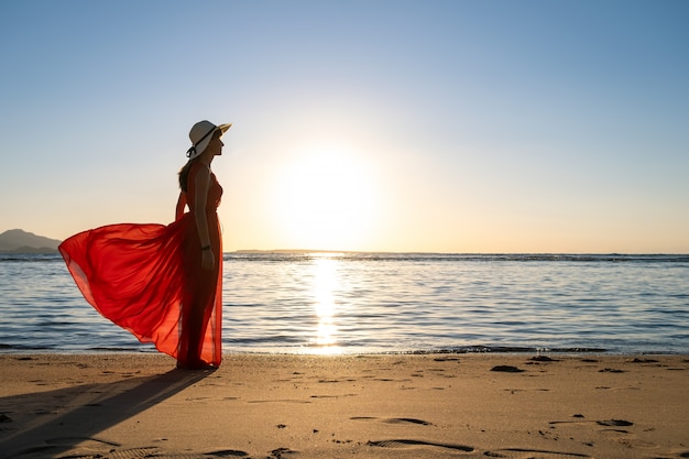 長い赤いドレスと夏の朝に昇る太陽の景色を楽しみながら海岸の砂浜に立っている麦わら帽子を着た若い女性