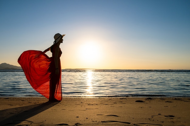Молодая женщина в длинном красном платье и соломенной шляпе, стоя на песчаном пляже на берегу моря