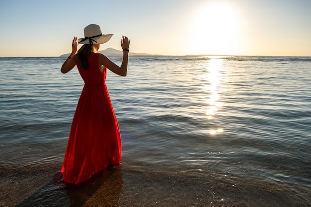 Foto giovane donna che indossa abito rosso lungo e cappello di paglia in piedi in acqua di mare in spiaggia godendo la vista del sol levante nella mattina di inizio estate.