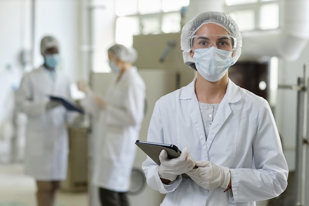 Young woman wearing mask and lab coat looking at camera in factory workshop