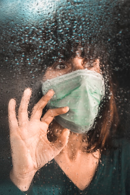 A young woman wearing a mask quarantined the Covid-19 pandemic at the window on a rainy day