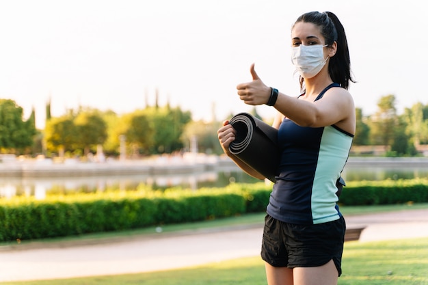 Young woman wearing a mask and sportswear gesturing to be fine with a mattress rolled up in her hand