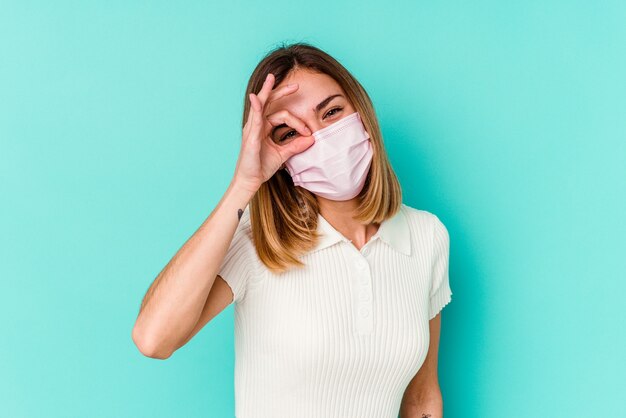 Young woman wearing a mask for virus isolated on blue excited keeping ok gesture on eye.