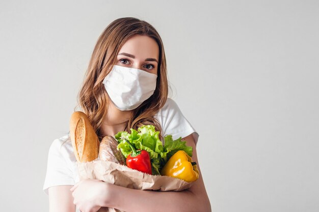 医療マスクを身に着けている若い女性は菜食主義の食糧、灰色の背景、宅配、コロノウイルス、検疫、ホームコンセプト、コピースペースを分離した上に分離された果物と野菜の紙袋を保持しています。