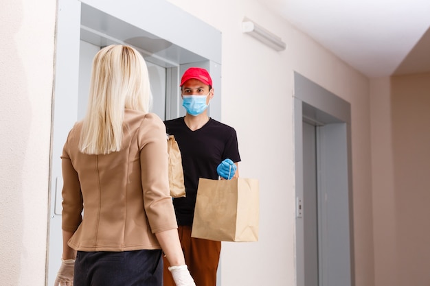 Young woman wearing medical mask receiving orderd food from the restaurant from delivery man indoors. Prevention of virus spread