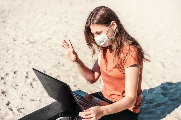 Giovane donna che indossa una maschera medica si siede sulla spiaggia con un computer portatile