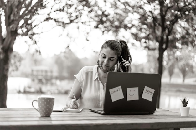 Giovane donna che indossa una maschera facciale aperta che chiama con lo smartphone mentre lavora con il laptop all'aperto. freelancer femminile sorridente che parla al cellulare e usa il pc al parco durante la fine dell'epidemia di covid19