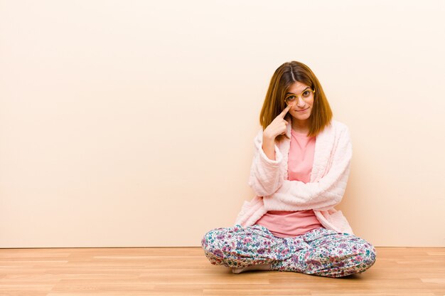 Young woman wearing pajamas sitting at home keeping an eye on you, not trusting, watching and staying alert and vigilant