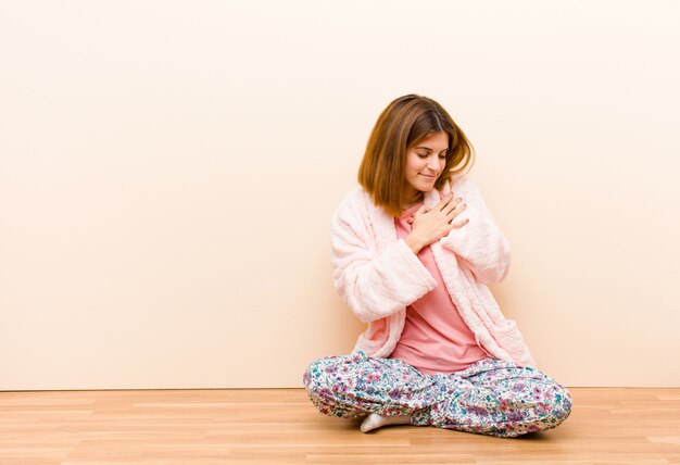 Young woman wearing pajamas sitting at home looking sad, hurt and heartbroken, holding both hands close to heart, crying and feeling depressed