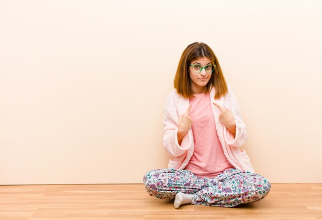 Young woman wearing pajamas sitting at home pointing to self with a confused and quizzical look, shocked and surprised to be chosen