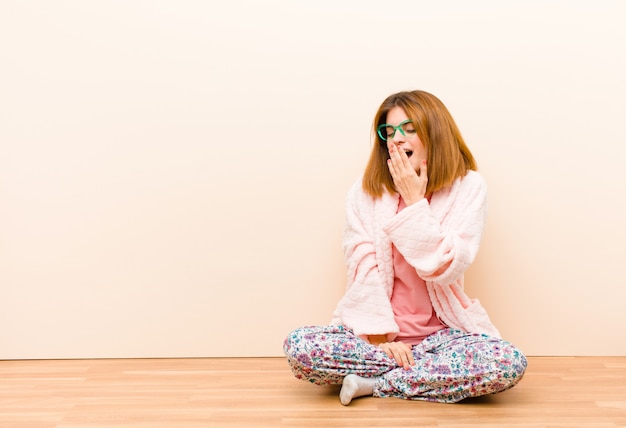 Young woman wearing pajamas sitting at home yawning lazily early in the morning waking and looking sleepy tired and bored