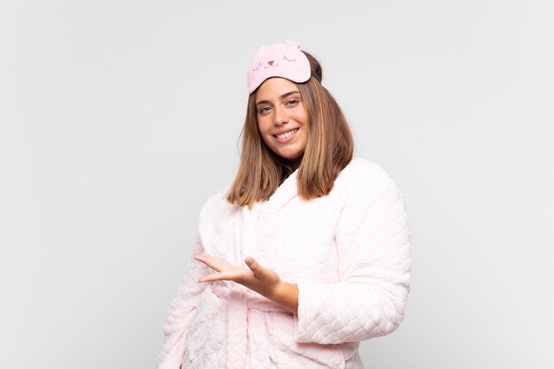 Young woman wearing pajamas, smiling cheerfully, feeling happy and showing a concept in copy space with palm of hand