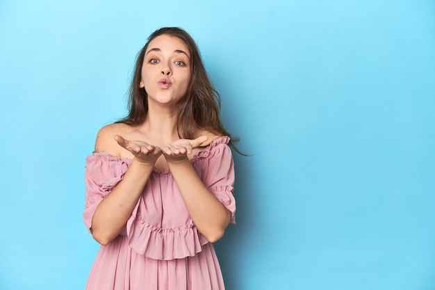 Young woman wearing a pink dress folding lips and holding palms to send air kiss