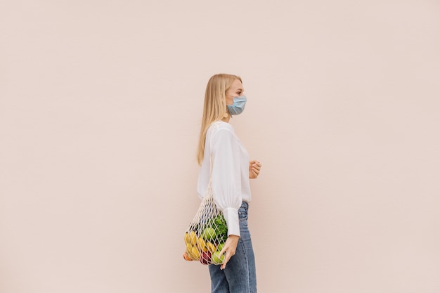 코로나바이러스 코비드-19 전염병 예방을 위해 보호용 안면 마스크를 쓴 젊은 여성이 과일이 든 끈 쇼핑백을 들고 있습니다. 에코 라이프 스타일. 의식적인 소비. 뉴 노멀. 복사 공간