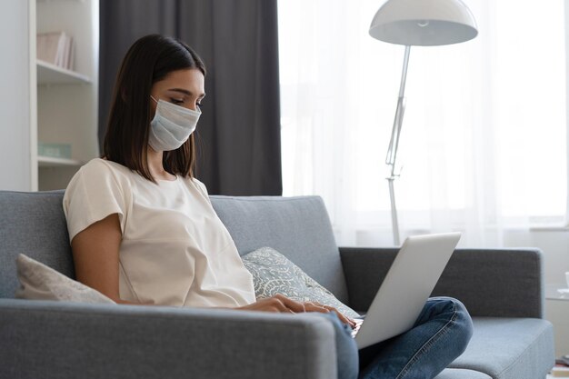 Photo young woman wearing protective mask in quarantine and using laptop while sitting on comfortable sofa