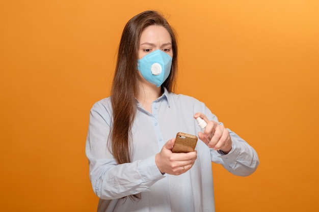 Young woman wearing protective mask and using spray on smartphone