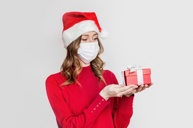 Молодая женщина в защитной медицинской маске и шляпе Санта держит красную подарочную коробку, изолированную на сером фоне студии. Новогодние подарки во время карантина по коронавирусу. Онлайн заказ