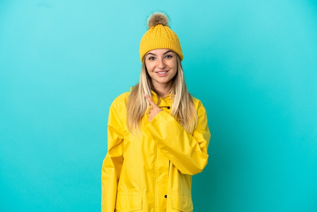 제품을 제시하기 위해 측면을 가리키는 격리된 파란색 배경 위에 방수 코트를 입은 젊은 여성