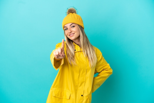 Young woman wearing a rainproof coat over isolated blue background showing and lifting a finger