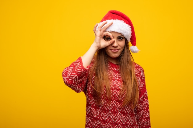 Young woman wearing santa hat confident doing ok gesture on eye