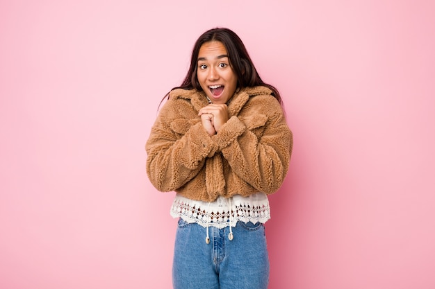 Young woman wearing a short sheepskin coat praying for luck, amazed and opening mouth looking to front