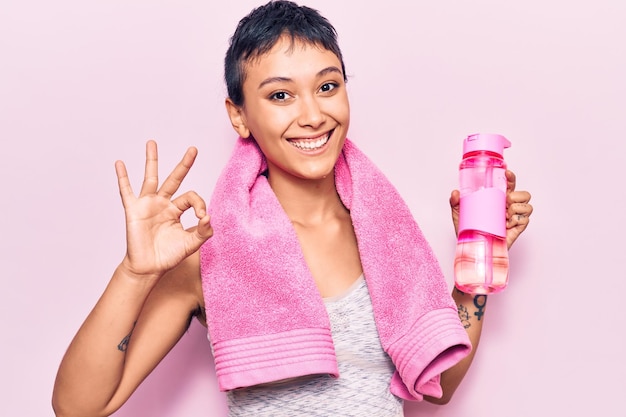 スポーツウェアを着た若い女性が水のボトルを持ち、笑顔のフレンドリーなジェスチャーで優れたシンボルを指でOKサインをしている