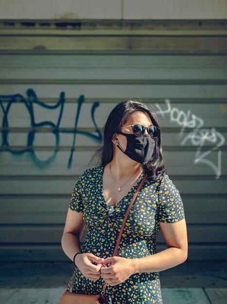 Young woman wearing sunglasses and mask while standing against wall