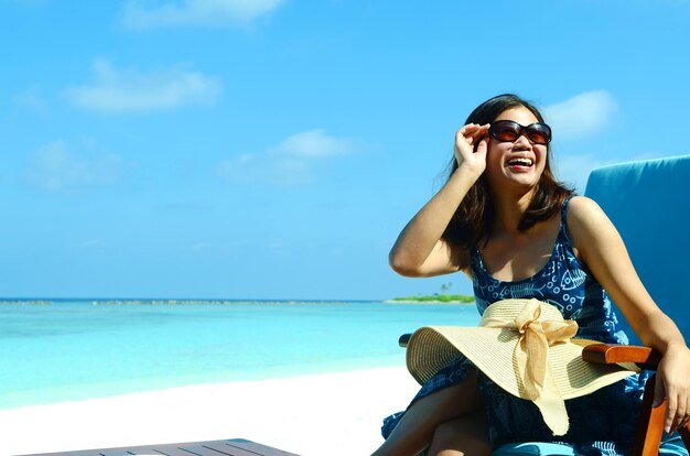 Foto giovane donna con gli occhiali da sole seduta sulla spiaggia contro il cielo