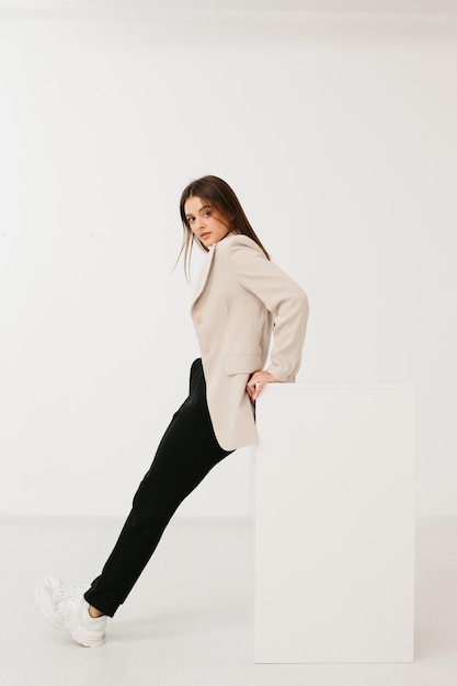 Young woman wearing trendy pastel blazer and pants with stylish sneakers sitting on cube against white background in studio