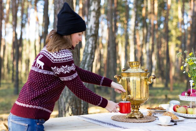 Young woman wearing warm jersey pouring some tea using vintage samovar at the forest