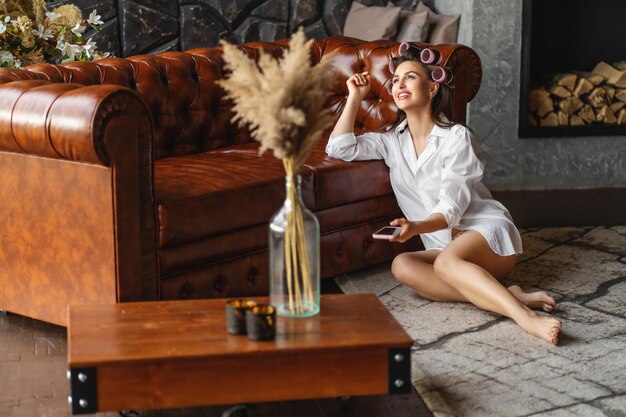 Young woman wearing a white shirt with curlers in her hair sitting near the sofa in and laughs after talking with friends on the phone