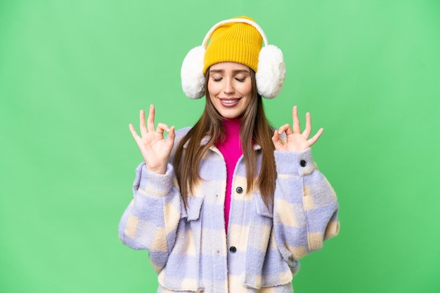Young woman wearing winter muffs over isolated chroma key background in zen pose