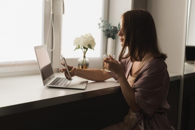 젊은 여성은 거실에 앉아 차 소셜 네트워크를 마시고 집에서 일하는 동안 노트북 컴퓨터에서 일하는 잠옷을 입는다