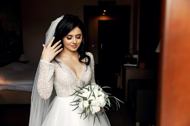 Young woman in a wedding dress drinking wine on the bed in a luxurious interior.
