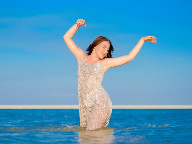 A young woman in a wet sundress enjoys splashing water while\
standing kneedeep in the sea