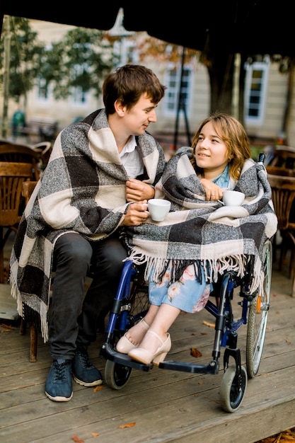 young woman on a wheelchair drinking coffee in a cafe outdoors with her husband and having a good time.