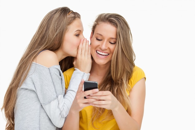 Giovane donna sussurrando alla sua amica che sta scrivendo sul suo telefono