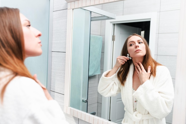 A young woman in a white bathrobe is standing in front of a mirror and doing a facial massageBright spacious bathroom