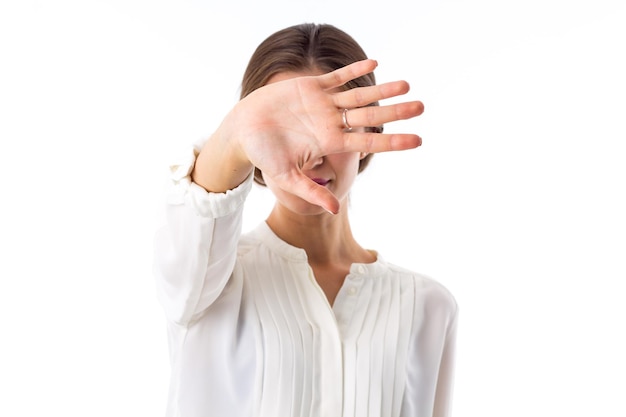 Young woman in white blouse hiding her face behind the hand on white background in studio