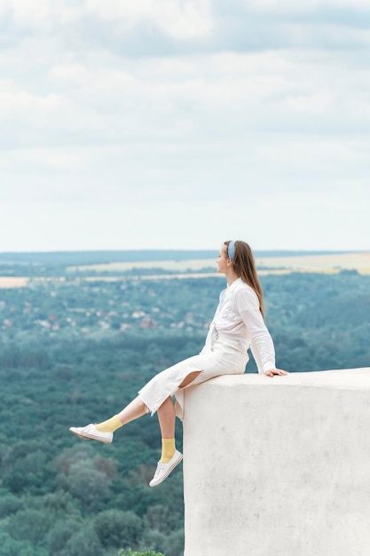 Young woman in white clothes sits on high observation deck with her legs hanging down Fulllength portrait Vertical frame