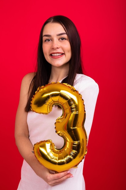 A young woman in a white dress holds a balloon in the form of the number three Model in the studio on a red background