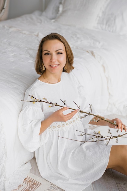 하얀 드레스를 입은 젊은 여성은 부활절 가지를 손에 들고 집 침대 옆에 앉아 있습니다.