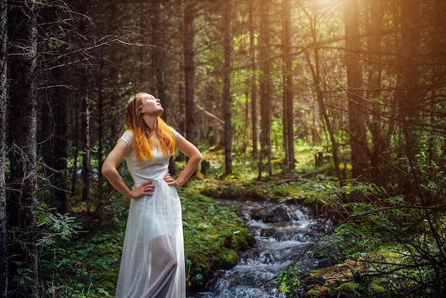 Foto giovane donna in abito bianco alzò il viso verso il sole bella ragazza sognante sullo sfondo della foresta verde e del ruscello di montagna